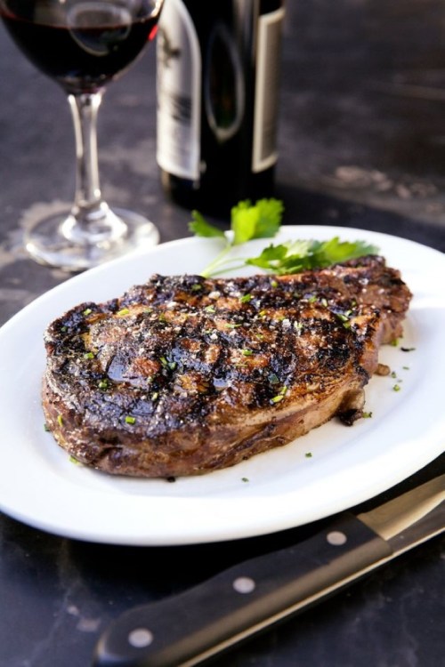 A perfectly grilled steak on a white plate, garnished with herbs, alongside a glass of red wine and a bottle.
