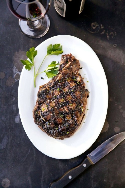 A grilled steak on a white plate, garnished with parsley, alongside a glass of red wine.