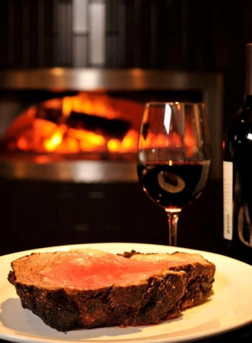 A slice of roast beef on a plate, accompanied by a glass of red wine, with a cozy fireplace in the background.