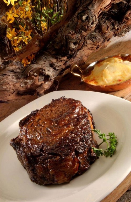 A beautifully cooked steak on a white plate, garnished with a sprig of thyme, with a side dish in the background.