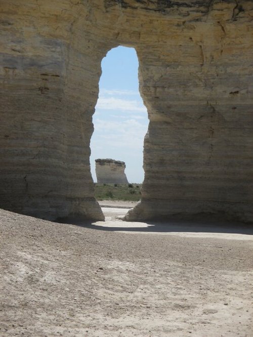 A natural rock formation with a large arch, framing a distant tower-like structure against a blue sky.