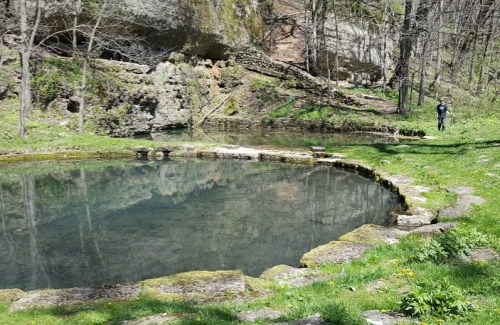 A serene pond surrounded by lush greenery and rocky terrain, with a person walking along the path nearby.