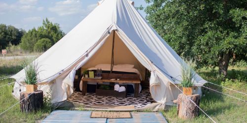 A spacious glamping tent with a bed and wooden table, set in a grassy area surrounded by trees.