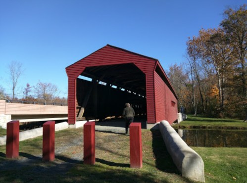 A person walks towards a red covered bridge surrounded by trees and a clear blue sky.