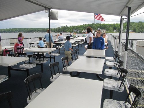 A spacious outdoor deck with tables and chairs, children and adults mingling, overlooking a lake and trees in the background.