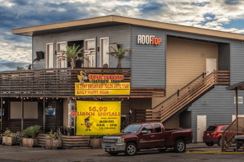 A two-story building with a rooftop sign, featuring colorful banners for breakfast tacos and lunch specials.