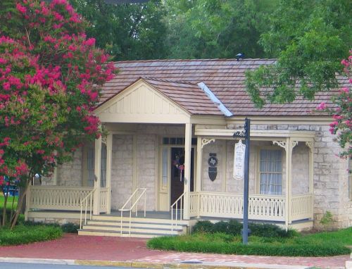 A charming yellow house with a porch, surrounded by pink flowering trees and a brick pathway.