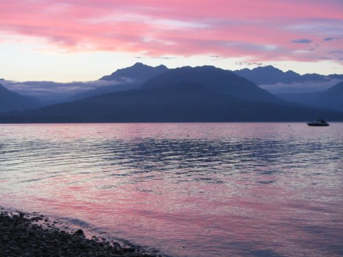 A serene lake at sunset, reflecting pink and purple hues, with mountains in the background and a small boat on the water.