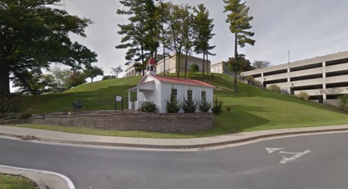 A small white building with a red roof sits on a grassy hill, surrounded by trees, near a parking garage.