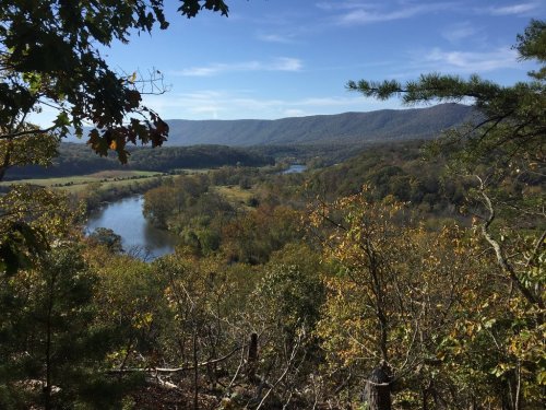A scenic view of a river winding through a valley, surrounded by autumn foliage and distant mountains under a clear blue sky.