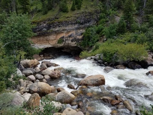 A rushing river flows over rocks near a cave, surrounded by lush greenery and steep, forested hills.