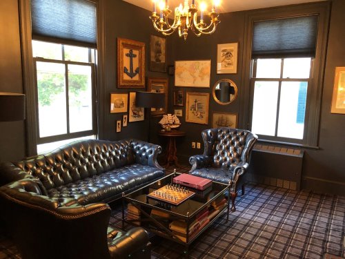 A cozy room with dark walls, leather sofas, a chess table, and nautical-themed decor, illuminated by a chandelier.