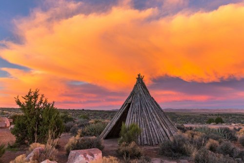 A wooden teepee stands in a desert landscape under a vibrant sunset sky with colorful clouds.