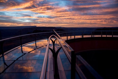 A glass walkway with railings overlooks a stunning sunset, reflecting vibrant colors in the sky and on the surface.
