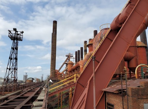 Industrial landscape featuring large rust-colored structures, smokestacks, and a railway track under a cloudy sky.
