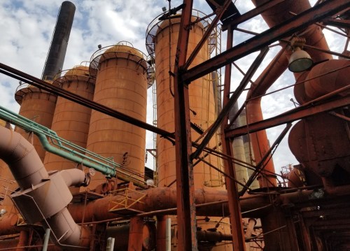 Industrial scene featuring large, rusted silos and pipes against a cloudy sky.