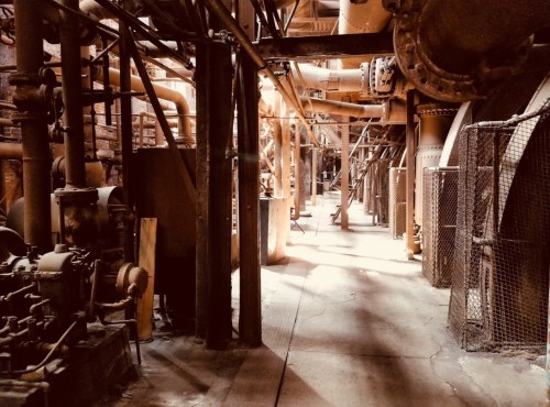 An industrial corridor filled with pipes and machinery, illuminated by soft light, creating a gritty atmosphere.