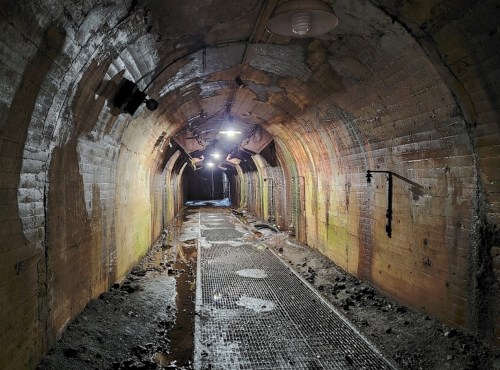 An empty, dimly lit tunnel with brick walls, wet floor, and metal grates, creating an eerie atmosphere.