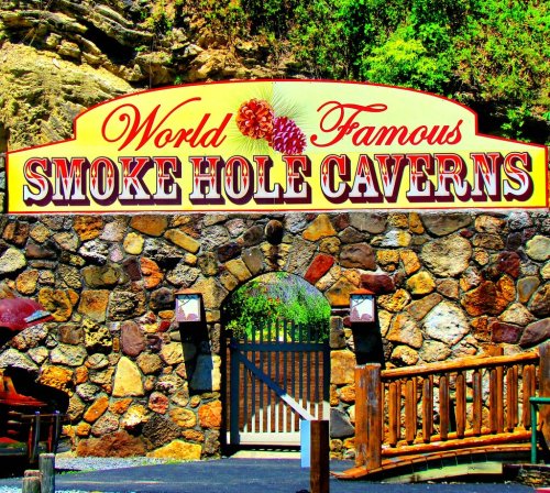 Sign for "World Famous Smoke Hole Caverns" at a stone entrance, surrounded by greenery and wooden railings.