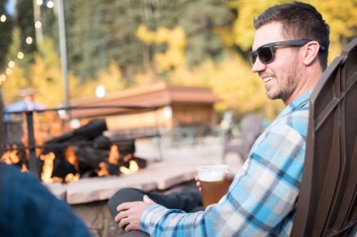 A man in sunglasses sits by a fire pit, enjoying a drink in a cozy outdoor setting surrounded by autumn trees.