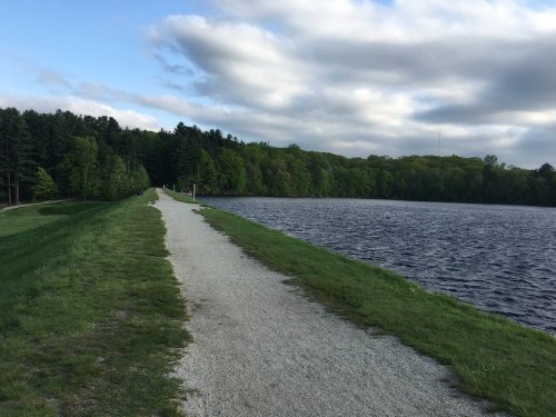 A gravel path alongside a calm lake, bordered by lush green trees under a partly cloudy sky.