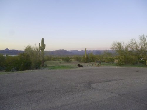 Desert landscape at dusk with cacti, mountains in the background, and a picnic table on a gravel area.