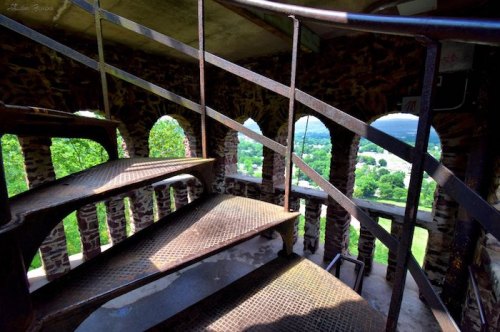 A spiral staircase inside a stone tower, with arched windows revealing a scenic view of greenery outside.