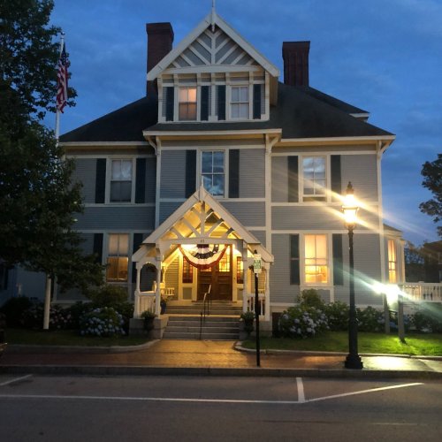 A large, historic house with a porch, lit windows, and American flags, set against a twilight sky.