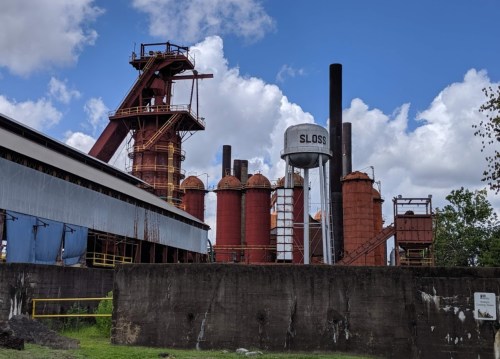 Industrial site featuring red silos, a water tower labeled "SLOSS," and a blue sky with scattered clouds.