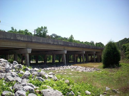 A concrete overpass supported by pillars, surrounded by greenery and rocky terrain under a clear blue sky.