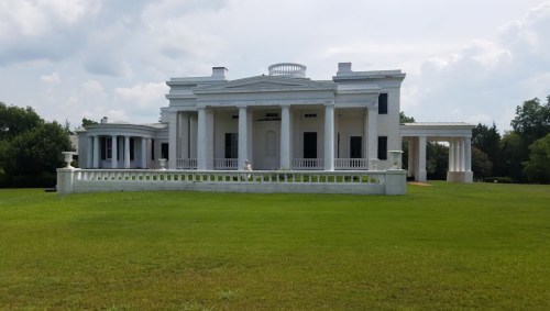 A large white mansion with columns, set on a green lawn under a cloudy sky.