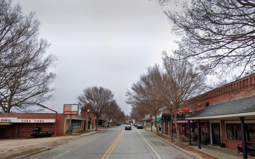 A quiet street lined with bare trees, shops, and a few cars under a cloudy sky.