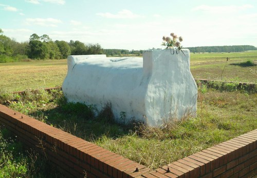 A large, white, abstract sculpture surrounded by a brick border, set in an open field with sparse vegetation.