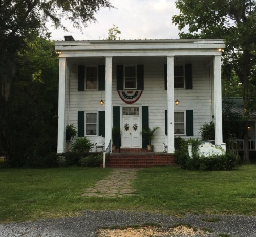 A two-story white house with green shutters, a front porch, and decorative wreaths, surrounded by greenery.