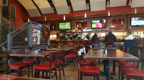 Interior of a restaurant with empty tables, bar area, and TVs showing sports, featuring red chairs and a warm atmosphere.