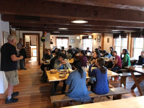 A group of people seated at long tables in a cozy dining area, listening to a speaker at the front.