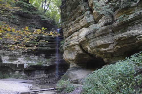 A serene waterfall cascading down rocky cliffs, surrounded by lush greenery and sandy ground.