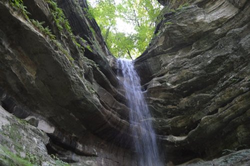 A waterfall cascades down rocky cliffs, surrounded by lush greenery and a bright sky above.