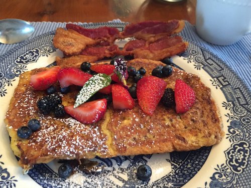A plate of French toast topped with strawberries, blueberries, and powdered sugar, alongside crispy bacon and a cup of coffee.
