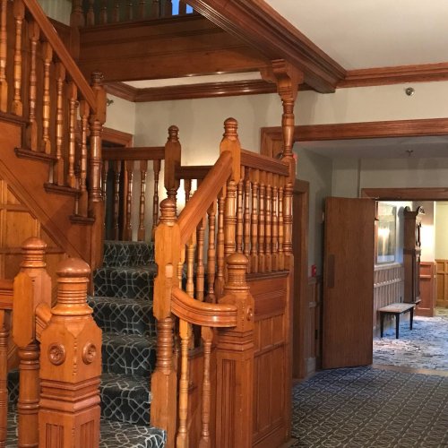 A wooden staircase with ornate railings, leading to a second floor, in a warmly lit hallway with patterned carpet.