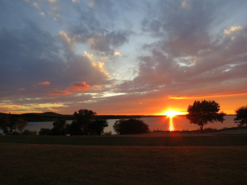 A vibrant sunset over a calm lake, with colorful clouds and silhouettes of trees along the shoreline.