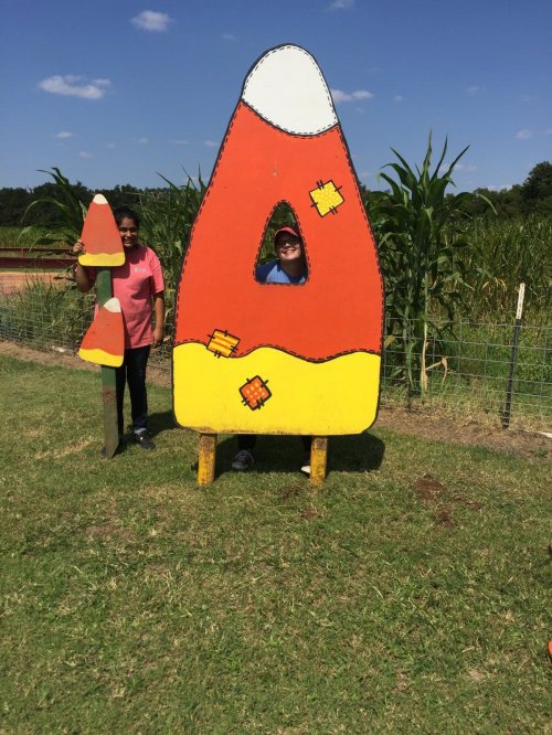 Two people stand behind a large candy corn cutout, smiling, with a cornfield in the background under a blue sky.