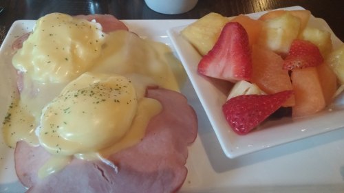 A plate of eggs Benedict with ham and hollandaise sauce, accompanied by a bowl of mixed fresh fruit.