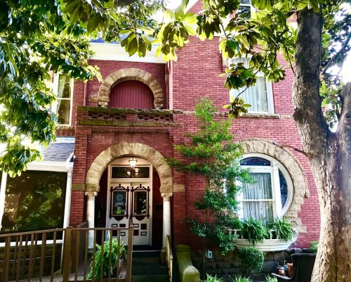 A charming red brick house with arched windows and a welcoming entrance, surrounded by lush greenery.