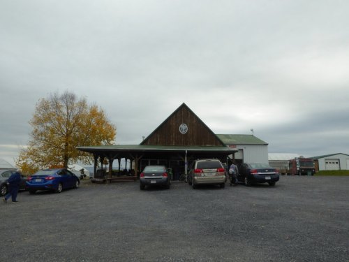 A rustic wooden building with a porch, surrounded by parked cars and a cloudy sky. Autumn trees are visible nearby.