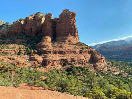 Red rock formations rise above a lush green landscape under a clear blue sky in a scenic outdoor setting.