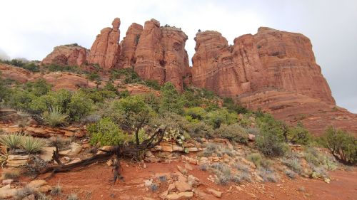 Red rock formations rise majestically against a cloudy sky, surrounded by green vegetation in a desert landscape.