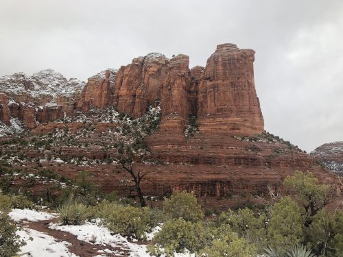 A snow-dusted red rock formation rises above green shrubs under a cloudy sky.