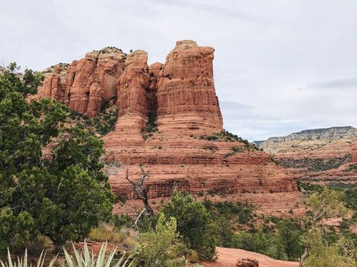 Red rock formations rise majestically against a cloudy sky, surrounded by green vegetation in a desert landscape.