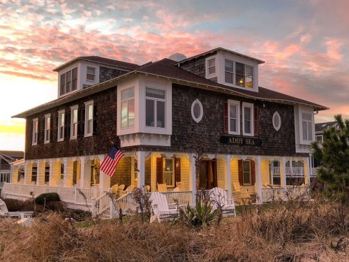 Historic seaside building with a sign reading "Addy Sea," featuring a porch and American flag, against a colorful sunset.